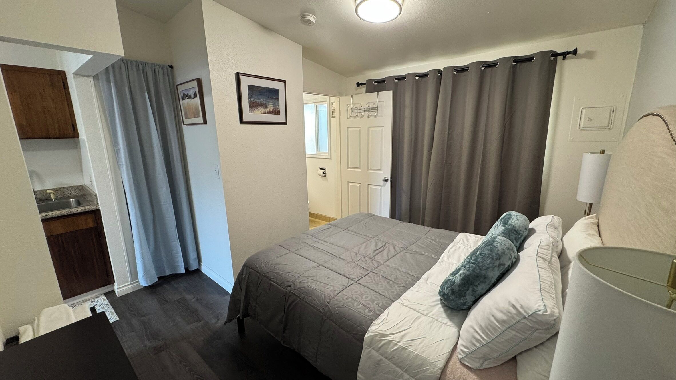 Un dormitorio con cama, almohadas, mesita de noche, lámpara, ventana con cortinas y puerta.