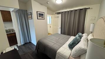 Un dormitorio con cama, almohadas, mesita de noche, lámpara, ventana con cortinas y puerta.