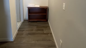 Een smalle gang met een houten vloer, een witte deur links en een klein tafeltje met een stapel witte handdoeken in het midden.