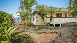 A house with a white porch, surrounded by tropical plants and a rocky garden. - Panoramic view of ocean.  Excellent location. Quiet neighborhood. Sleep 10 (Kailua-Kona)