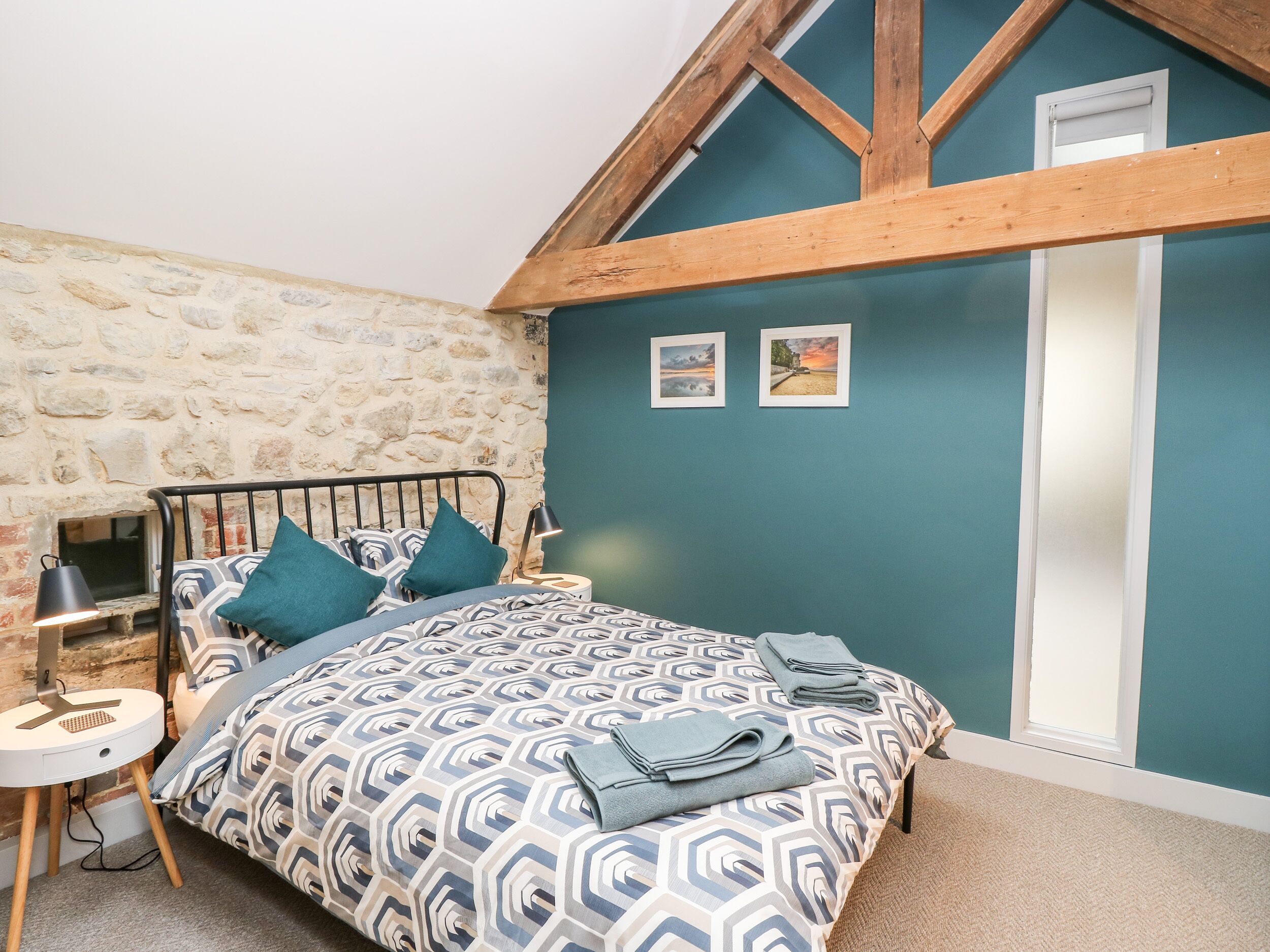 A bedroom with a stone wall, a bed with patterned bedding, a nightstand, a lamp, and two framed pictures on the wall.