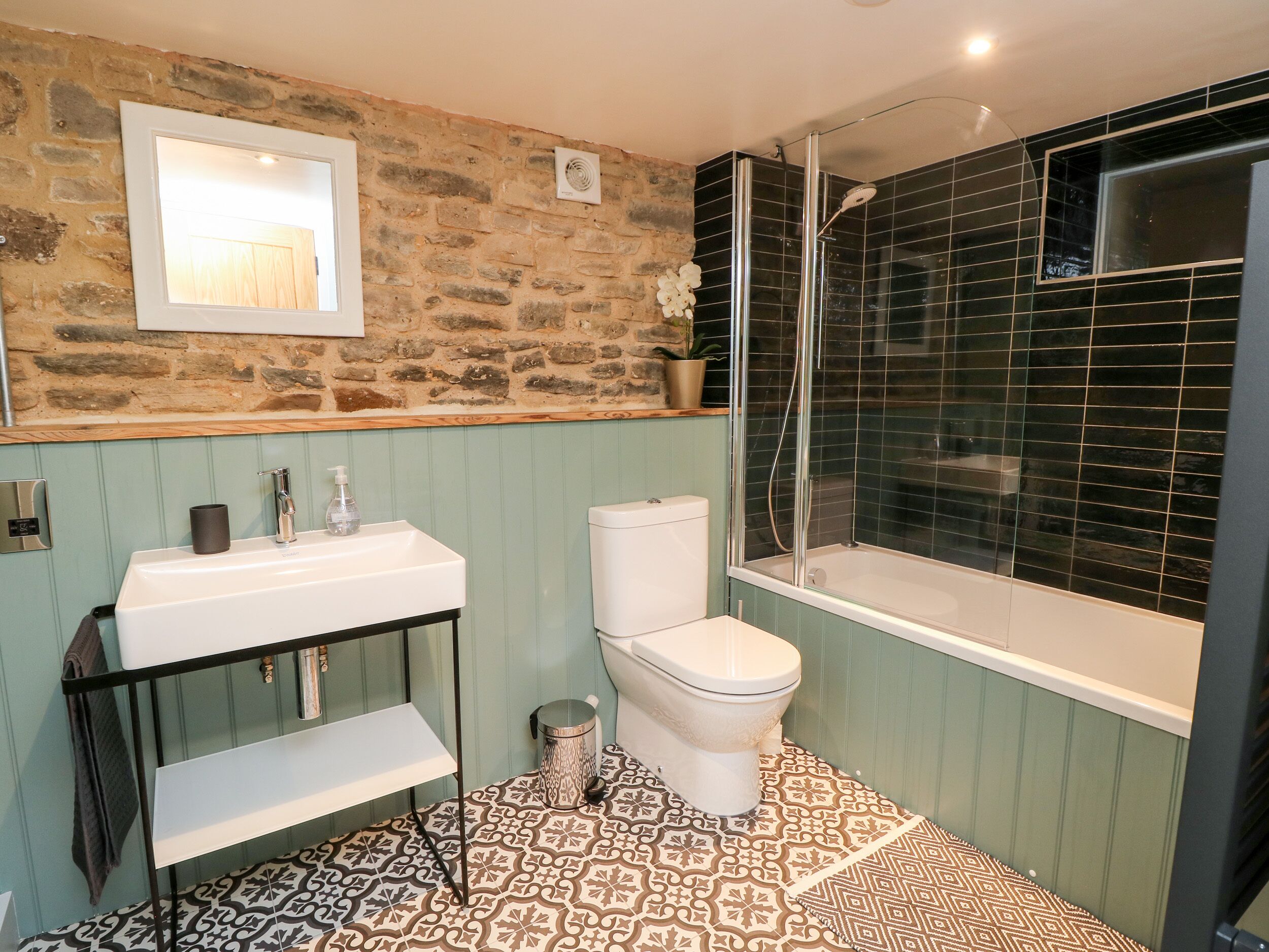 A bathroom with a stone wall, a white toilet, a sink with a mirror, and a shower area with black tiles.