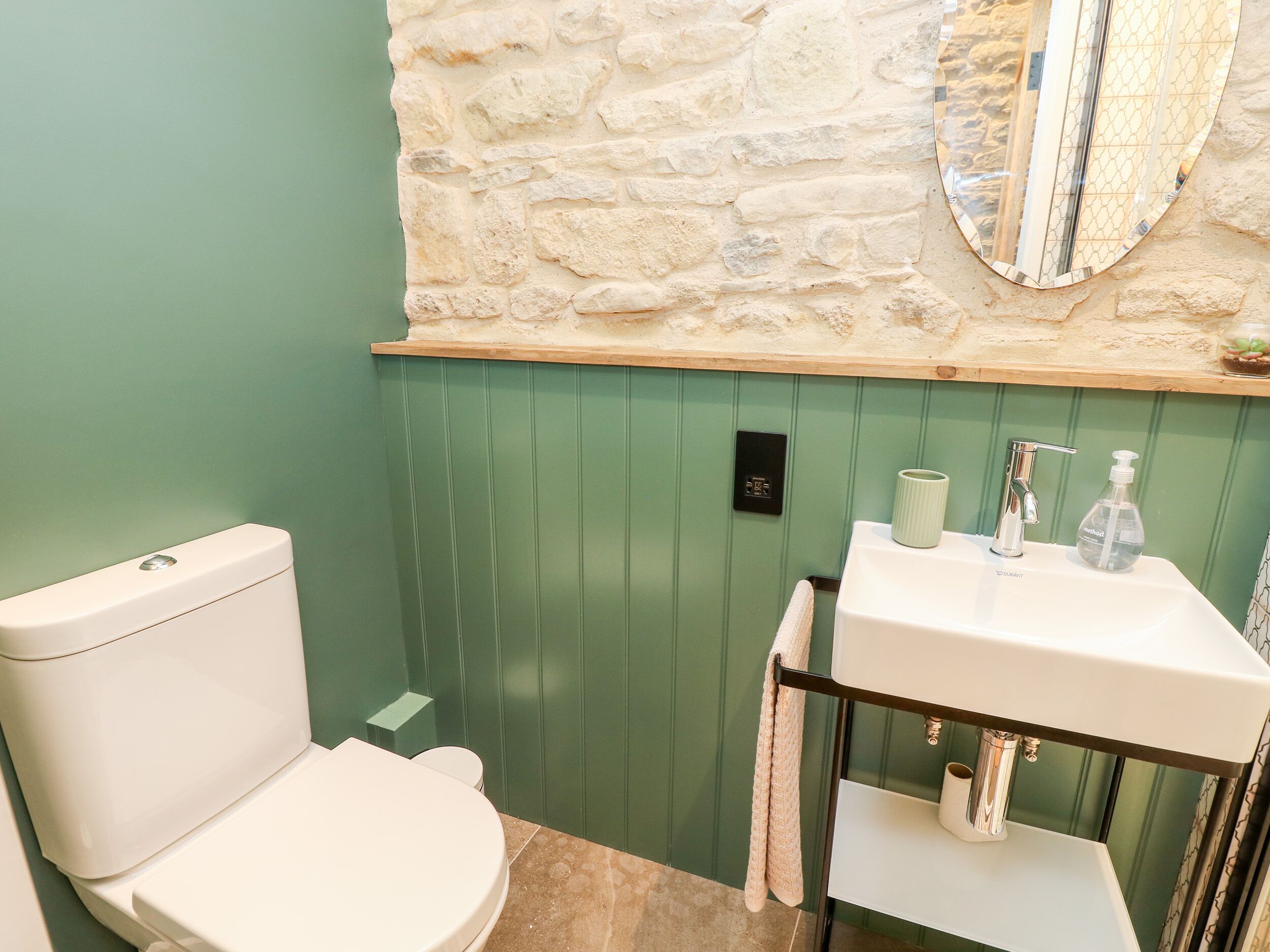 Une salle de bain avec des toilettes blanches, un lavabo avec un miroir et un mur vert à l’aspect pierre.