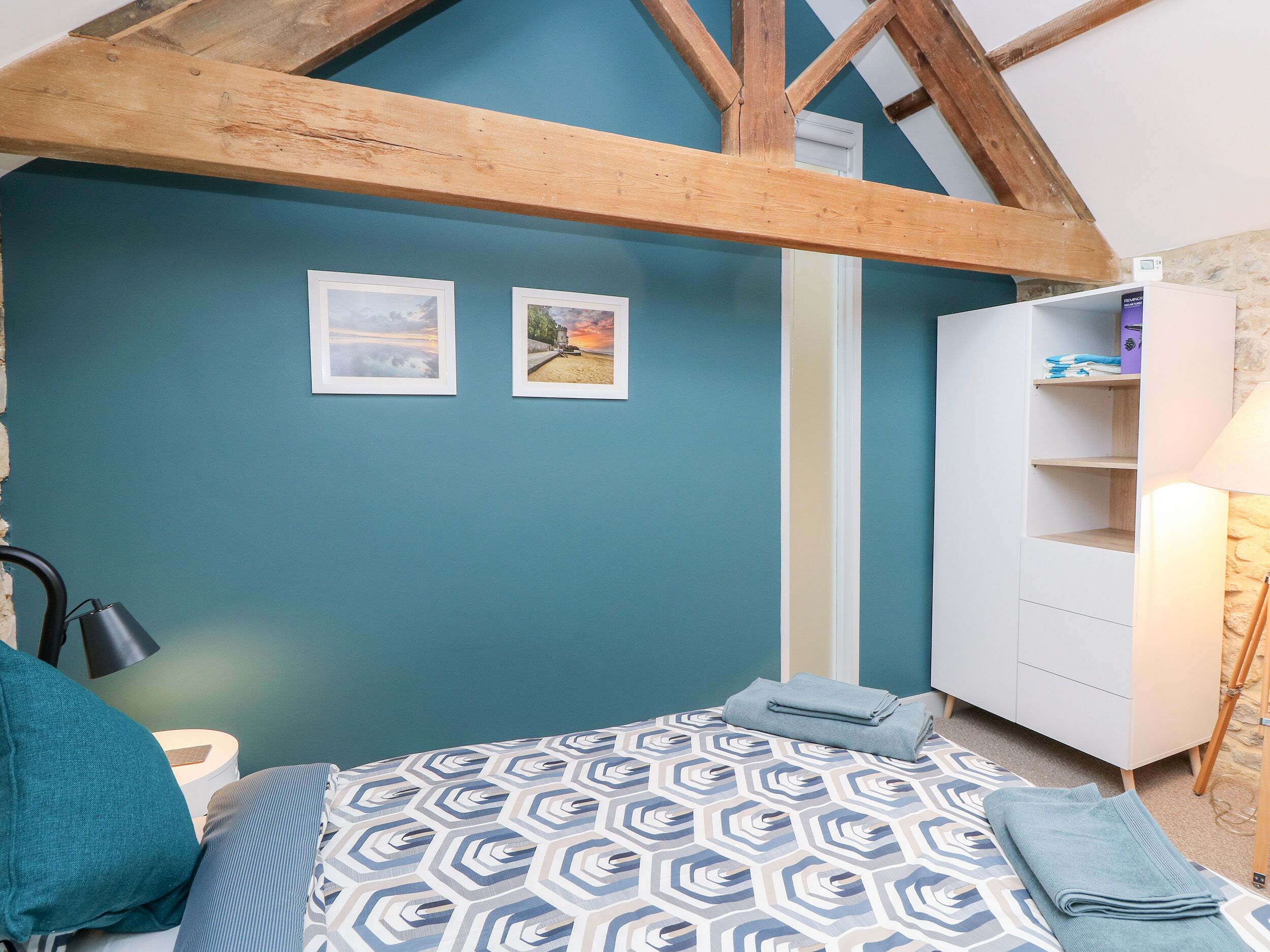 Une chambre avec un plafond en pente, un lit recouvert d’une literie à motifs, deux tableaux encadrés accrochés au mur et une armoire blanche.