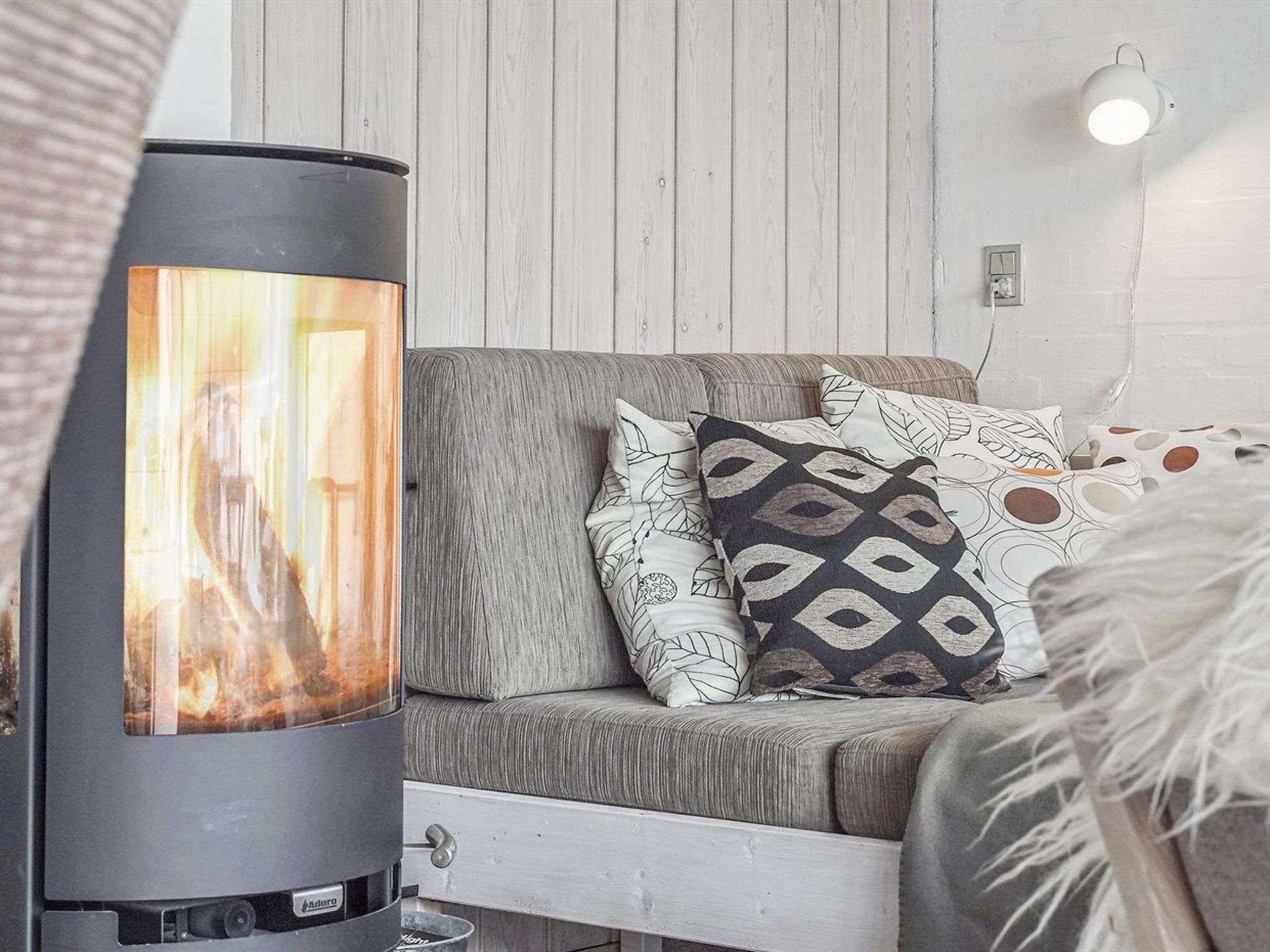 Una sala de estar acogedora con una chimenea moderna, un sofá gris con cojines estampados y una pared blanca con una lámpara.