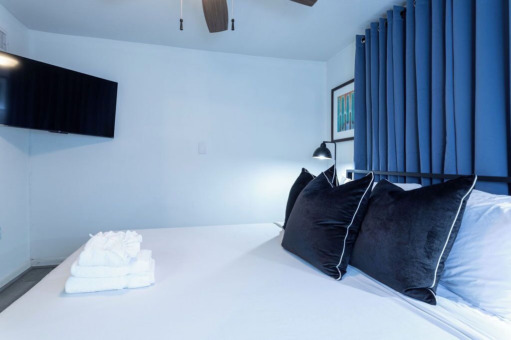 Une chambre d’hôtel moderne dotée d’un téléviseur à écran plat fixé au mur, d’un lit avec du linge de lit blanc et des oreillers noirs, d’une table de chevet avec une lampe et d’une grande fenêtre avec des rideaux bleus.