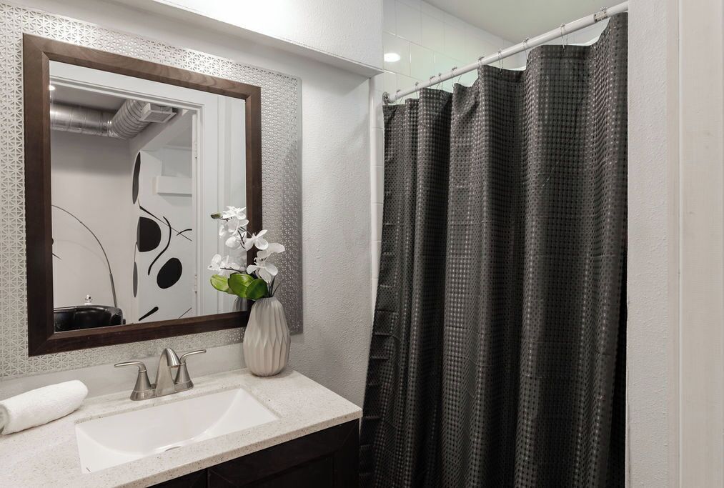 Um banheiro com pia branca, espelho com moldura decorativa, um vaso com flores brancas e um chuveiro com cortina quadriculada preta e branca.