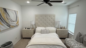 Une chambre à coucher avec un grand lit, deux tables de chevet, un ventilateur de plafond, un tableau accroché au mur et une fenêtre avec des stores.