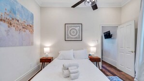 A hotel room with a bed, two bedside tables, a ceiling fan, a painting, a framed artwork, and a door.