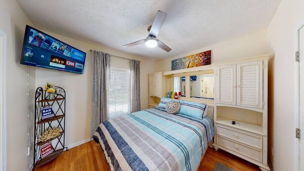 A bedroom with a bed, a ceiling fan, a TV mounted on the wall, and a shelf with items.