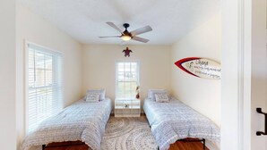 A bedroom with two single beds, a ceiling fan, a window with blinds, and a wall decoration.