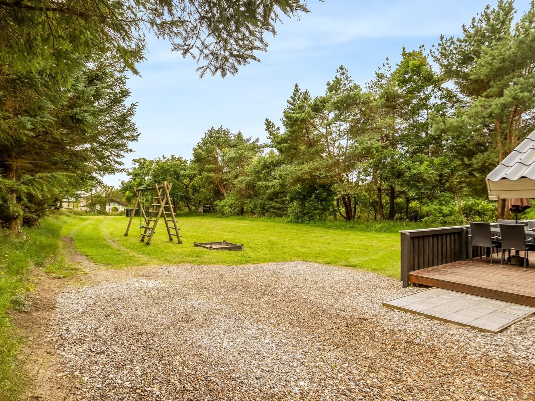 Un jardin avec un chemin de gravier, une balançoire en bois, une terrasse avec des meubles d’extérieur et une maison en arrière-plan.