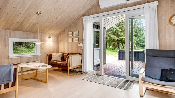 A cozy cabin interior with a wooden ceiling, a leather sofa, a dining table, and a large sliding glass door leading to an outdoor deck.