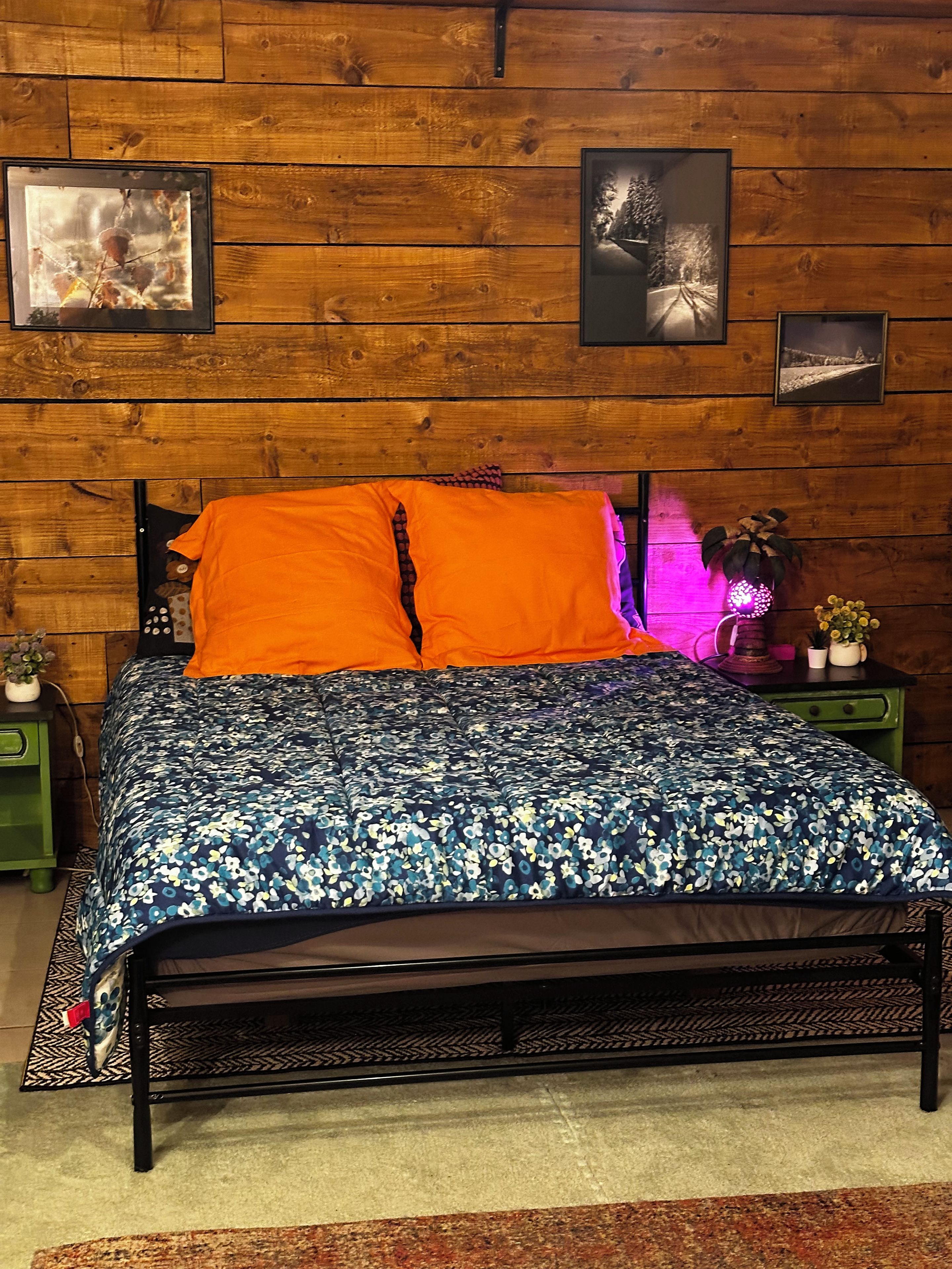 Ein Bett mit einem blumigen Überwurf, zwei Kissen und einem Kopfteil, vor einer Holzwand mit gerahmten Bildern.