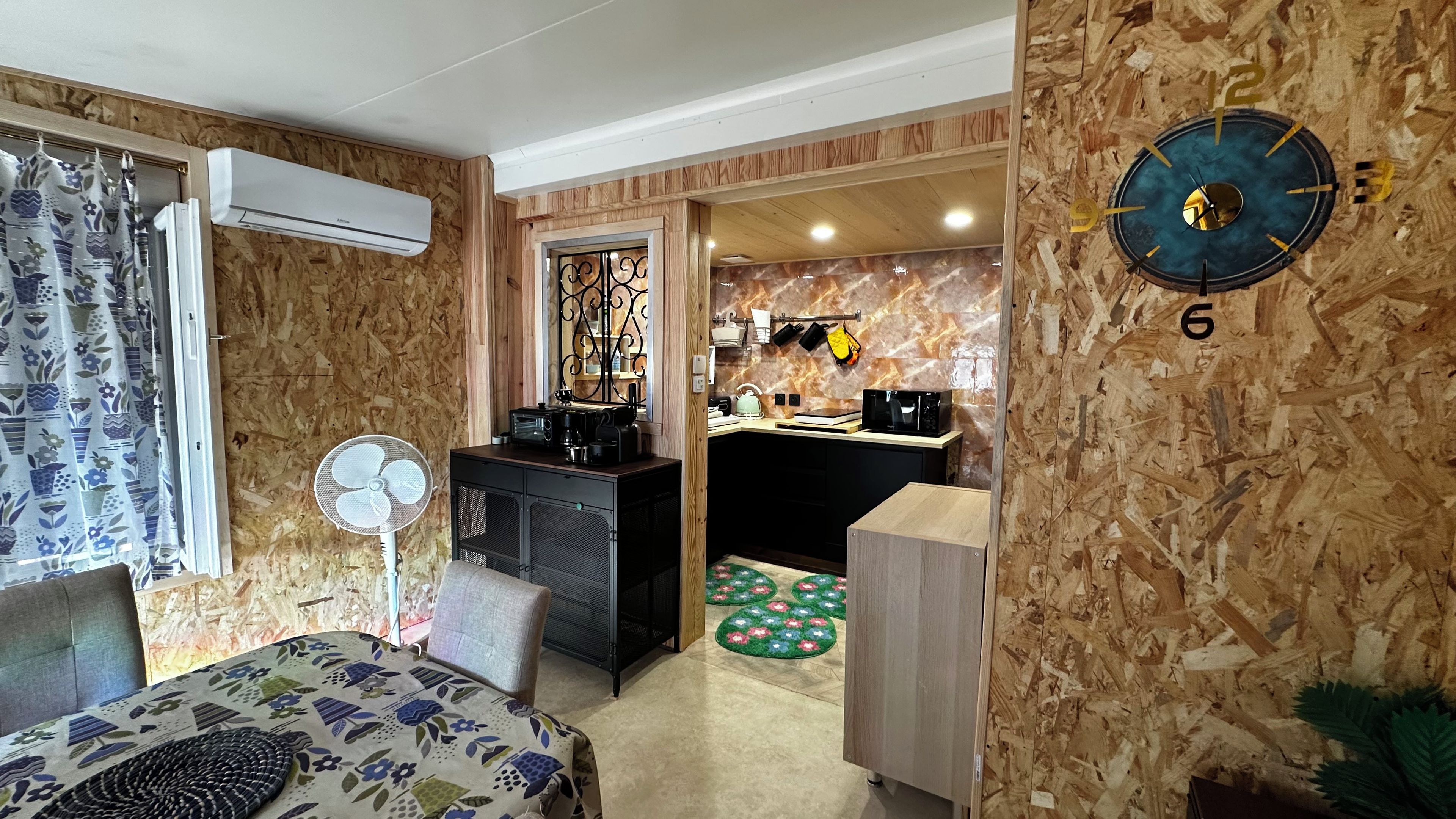 Quarto de hotel com cama, escrivaninha, ventilador, cozinha compacta e relógio na parede.