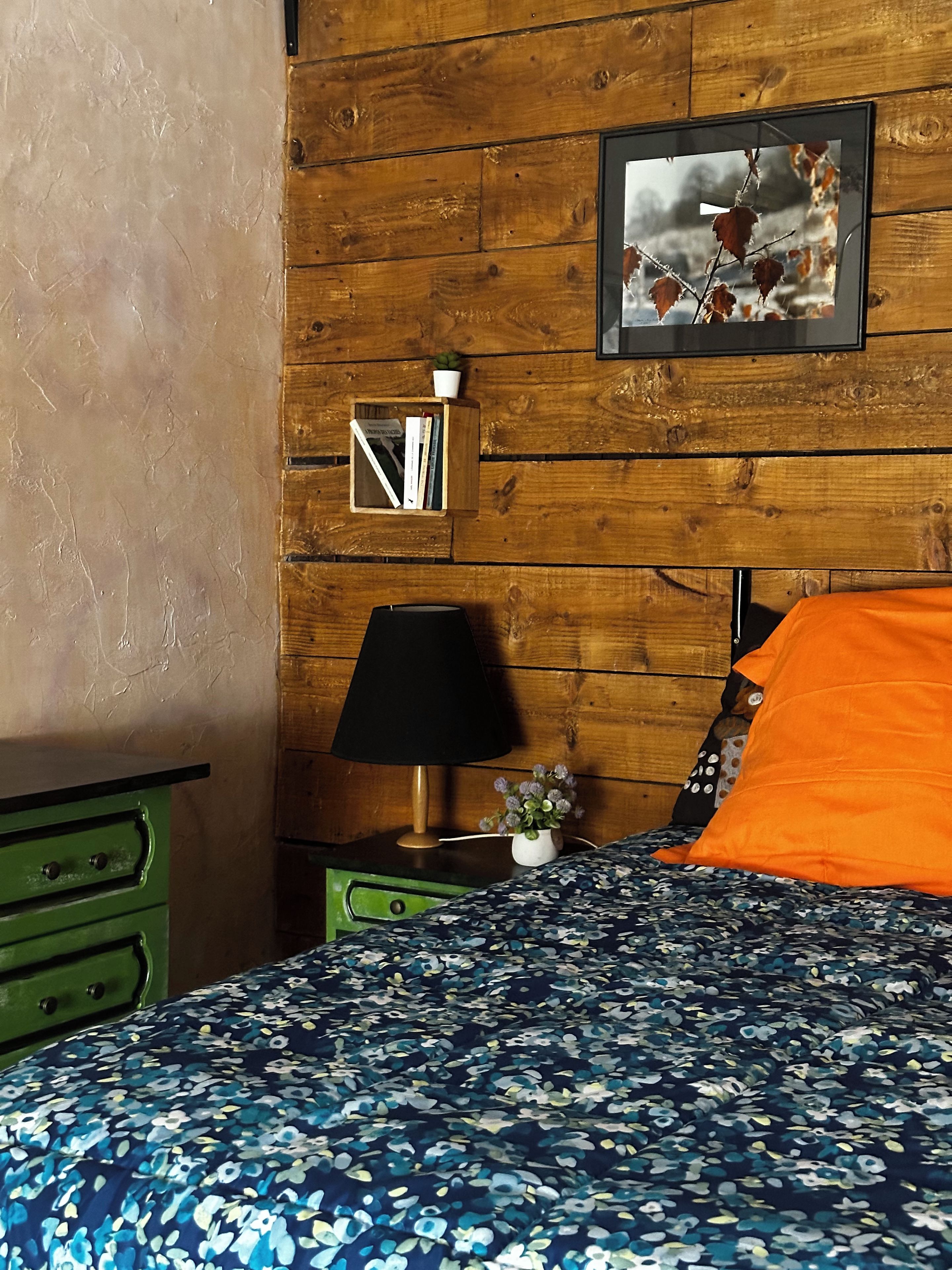 Une chambre avec un mur en bois, un lit avec un oreiller orange, une table de chevet verte, une lampe et un tableau encadré.