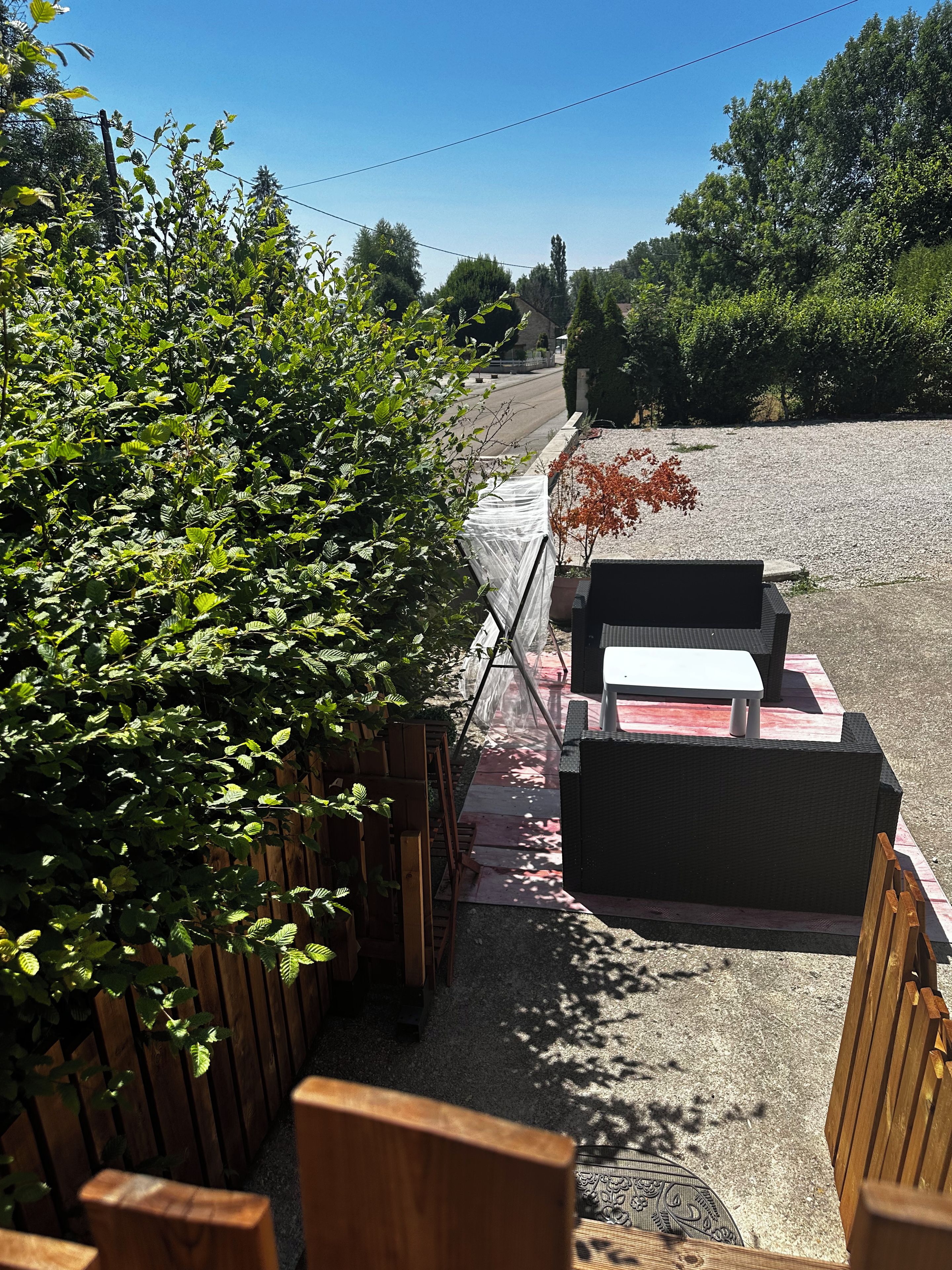 Un espace extérieur aménagé avec des bancs en bois et une table, entouré de verdure et bordé d’un chemin de gravier.