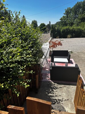 An outdoor seating area with wooden benches and a table, surrounded by greenery and a gravel path.