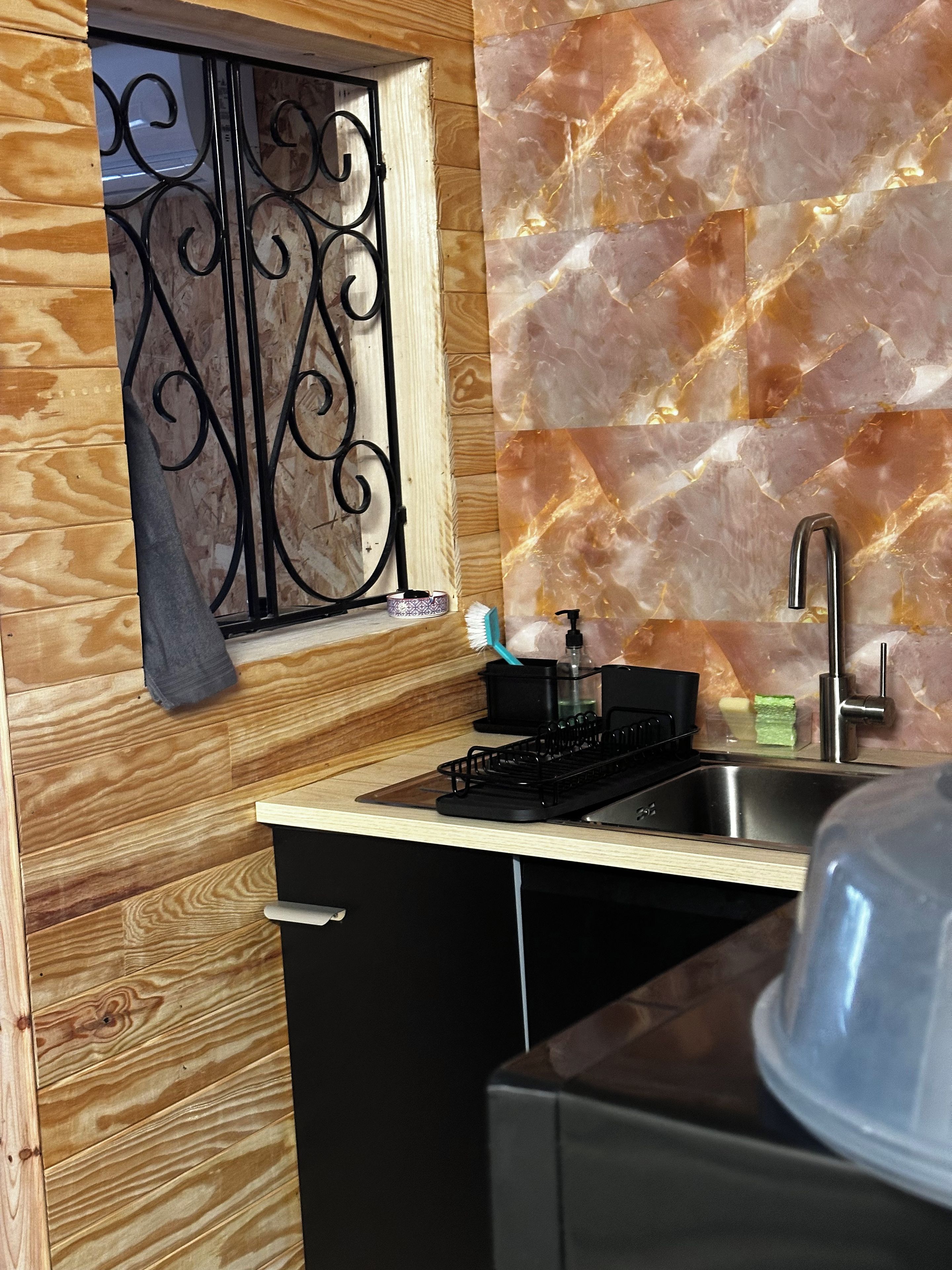 Een compacte keuken met een houten spatwand, een raam met decoratieve ijzeren tralies, een spoelbak en een kookplaat.