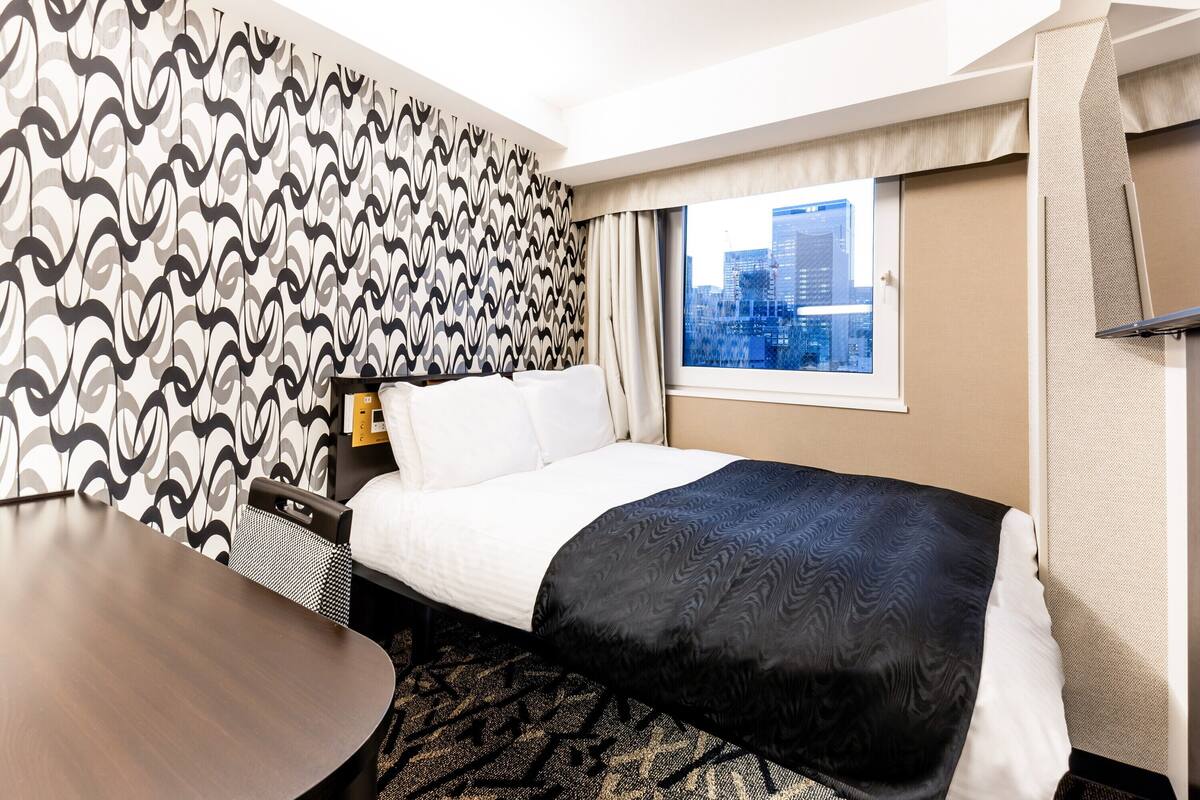 A hotel room with a bed, a desk, and a window with a city view.