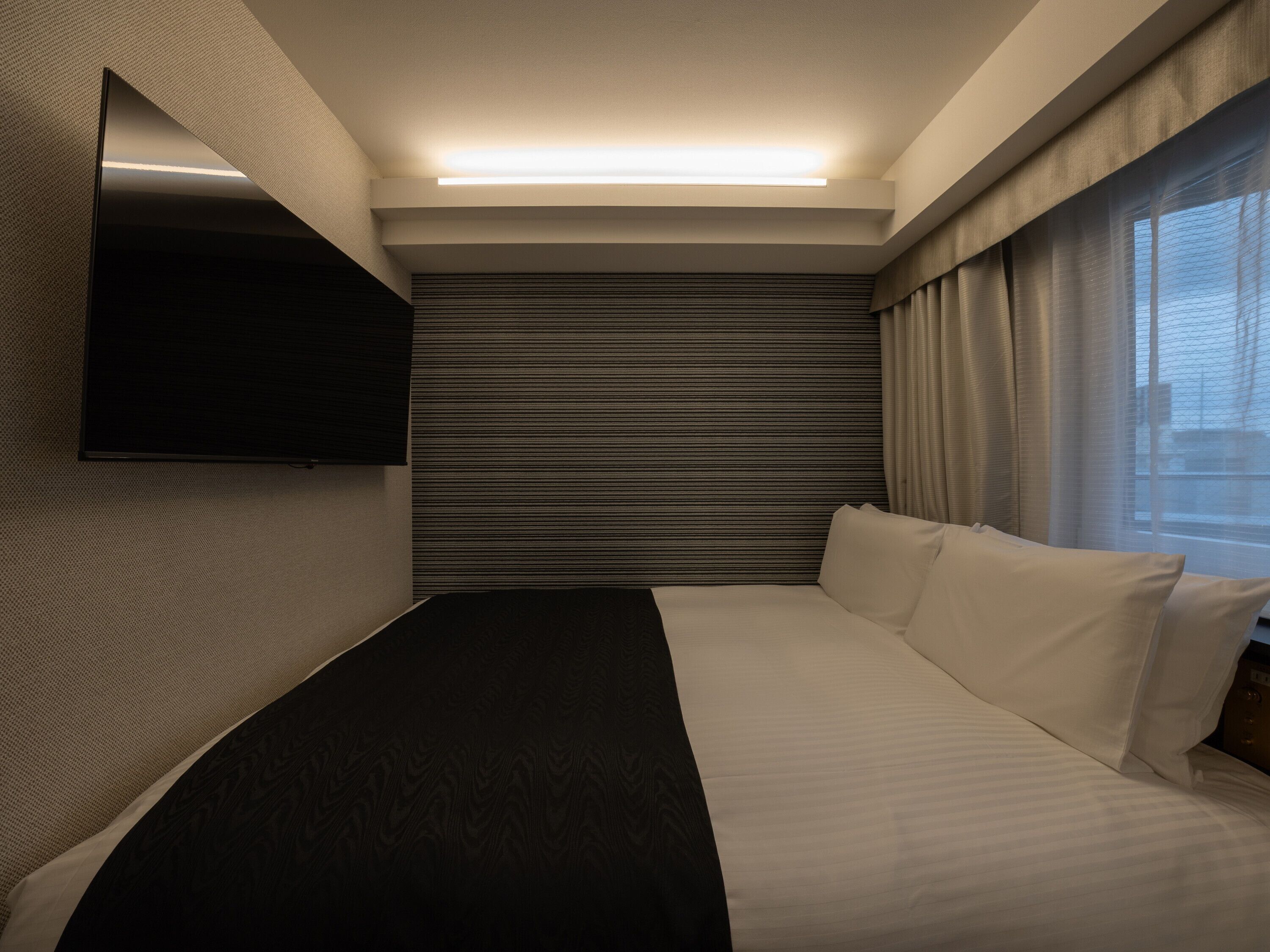 A modern hotel room with a large bed, a flat-screen TV mounted on the wall, and a window with curtains.