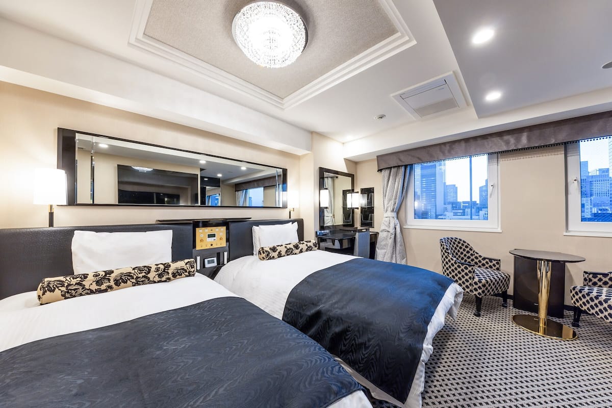 A hotel room with two beds, a desk, a chair, a small table, and a large window with a city view.