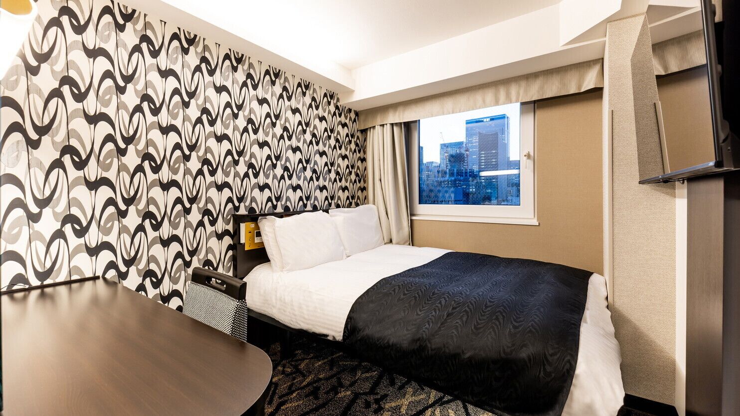 A hotel room with a bed, a desk, a window with a city view, and patterned wallpaper.