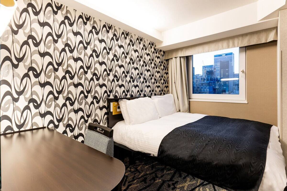 A hotel room with a bed, a desk, a window with a city view, and patterned wallpaper.