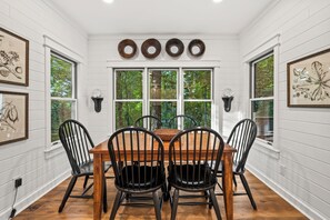 A dining room with a wooden table and four chairs, surrounded by white paneled walls and large windows.