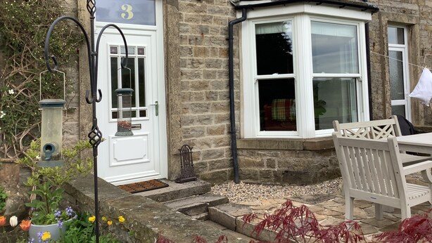 A stone building with a white door marked with the number 3, a bay window, and a bench outside.