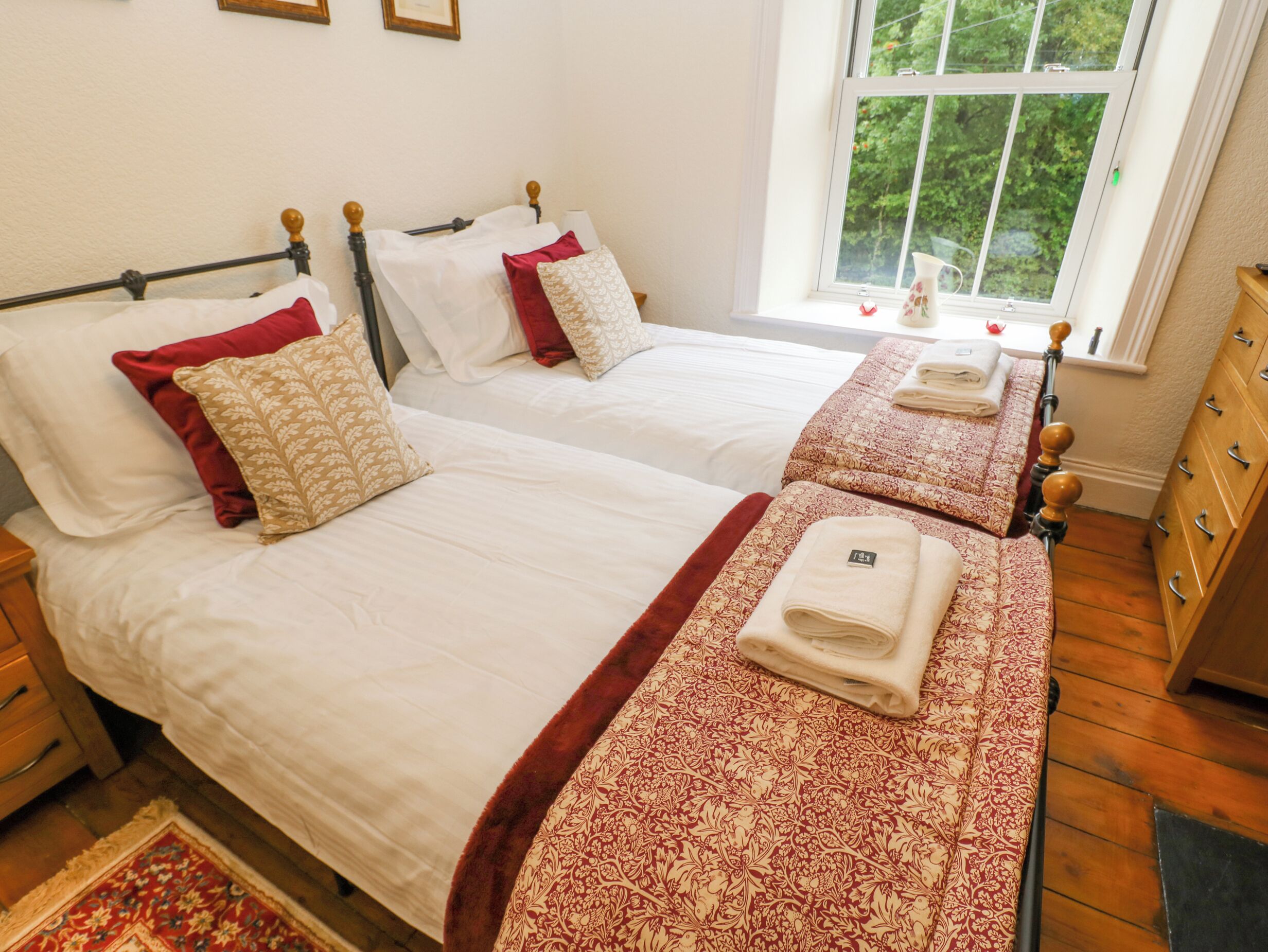 A hotel room with two beds, each with white linens and red and beige patterned pillows. There is a window with a view of greenery outside.