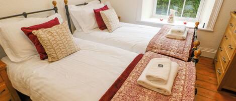 A hotel room with two beds, each with white linens and red and beige patterned pillows. There is a window with a view of greenery outside.