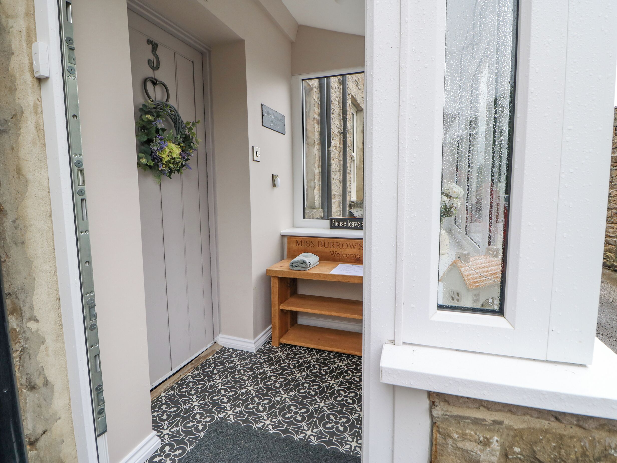 A narrow hallway with a door, a small bench, a welcome sign, and a mirror reflecting the outside.