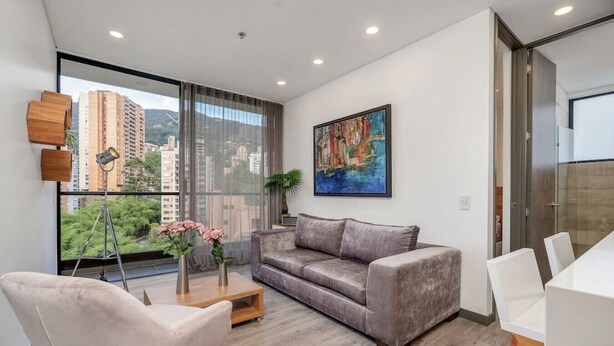 A modern living room with a grey sofa, a wooden coffee table, and a white chair. There is a painting on the wall and a balcony with a view of buildings and greenery.