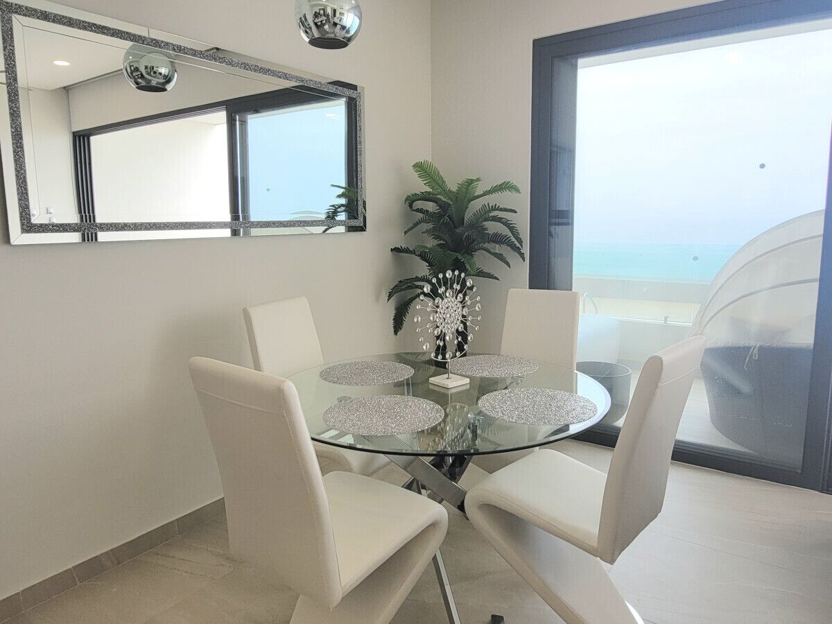 Un comedor con una mesa de vidrio rodeada de sillas, un centro decorativo, un espejo grande y vistas al exterior.