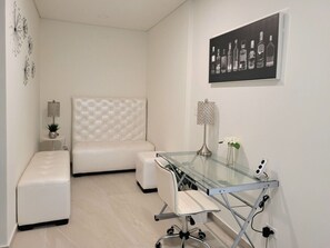 Une chambre moderne avec un lit à tête de lit capitonnée, un bureau en verre avec une lampe et une œuvre d’art fixée au mur représentant des bouteilles de spiritueux.