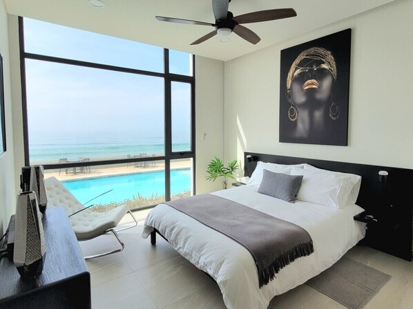 Une chambre avec une grande fenêtre donnant sur une piscine et l’océan, un lit avec une literie blanche et grise, un ventilateur de plafond et un tableau accroché au mur.