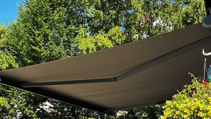 An outdoor seating area with plastic chairs and tables, shaded by a large canopy, surrounded by greenery and trees.