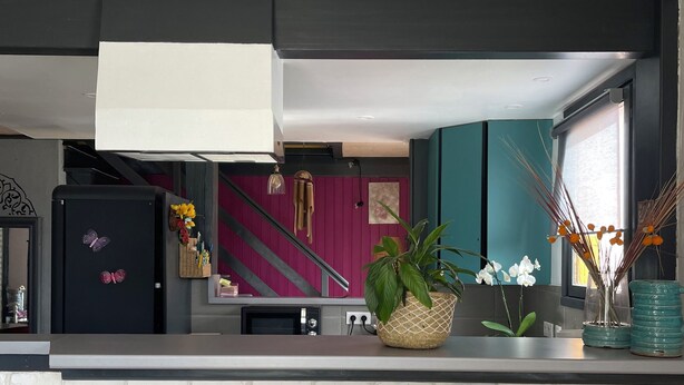 A modern kitchen with a white brick backsplash, two black bar stools, a black refrigerator with a butterfly decoration, a potted plant, and a window with a view of the outside.