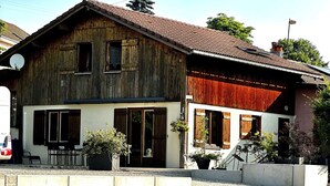 A two-story wooden house with a red roof, a front porch, and a small garden area with potted plants.