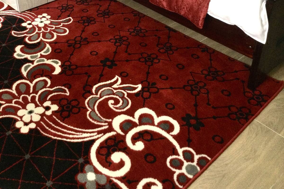 A neatly made bed with a red and white bedspread, a matching red throw blanket, and two red pillows. There is a small bedside table with a bottle of water on it. The room has a red patterned rug with floral designs.