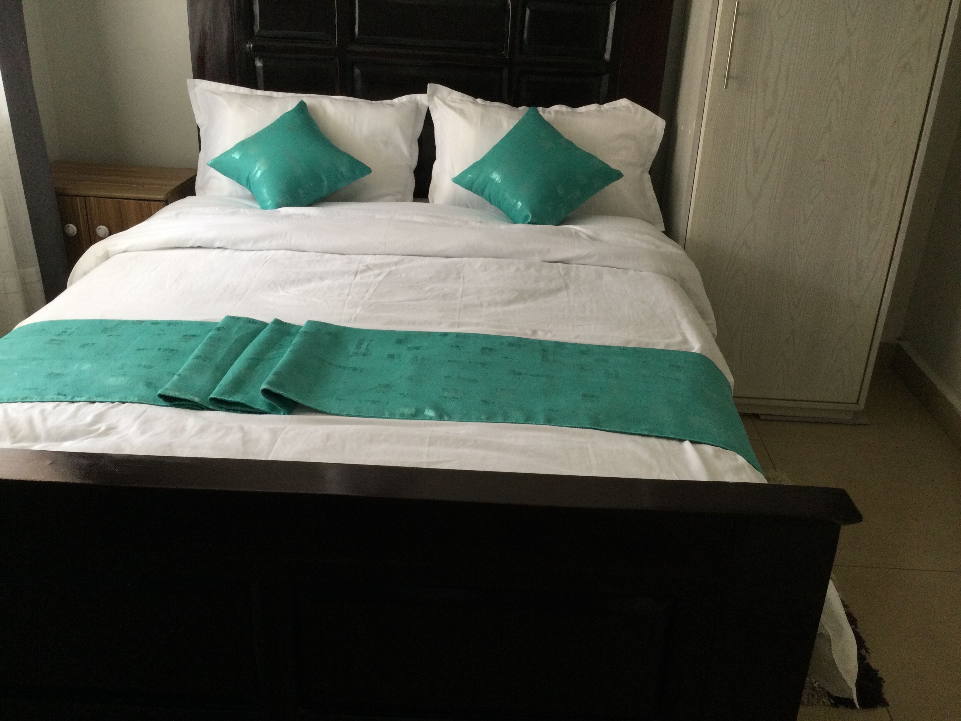 A neatly made bed with white bedding and teal pillows, a teal bedspread, and a dark wooden headboard.