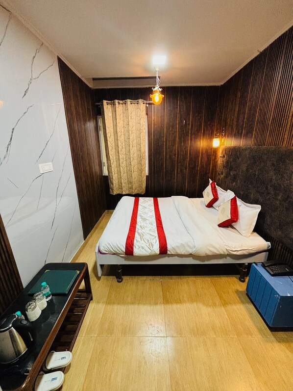 Un dormitorio con cama, suelo de madera, una mesita con hervidor y una pared con lámpara.