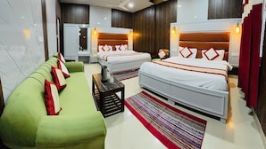 A hotel room with two beds, a green sofa, a small table, and a red and white striped rug. - HOTEL AMAR (Dehradun)