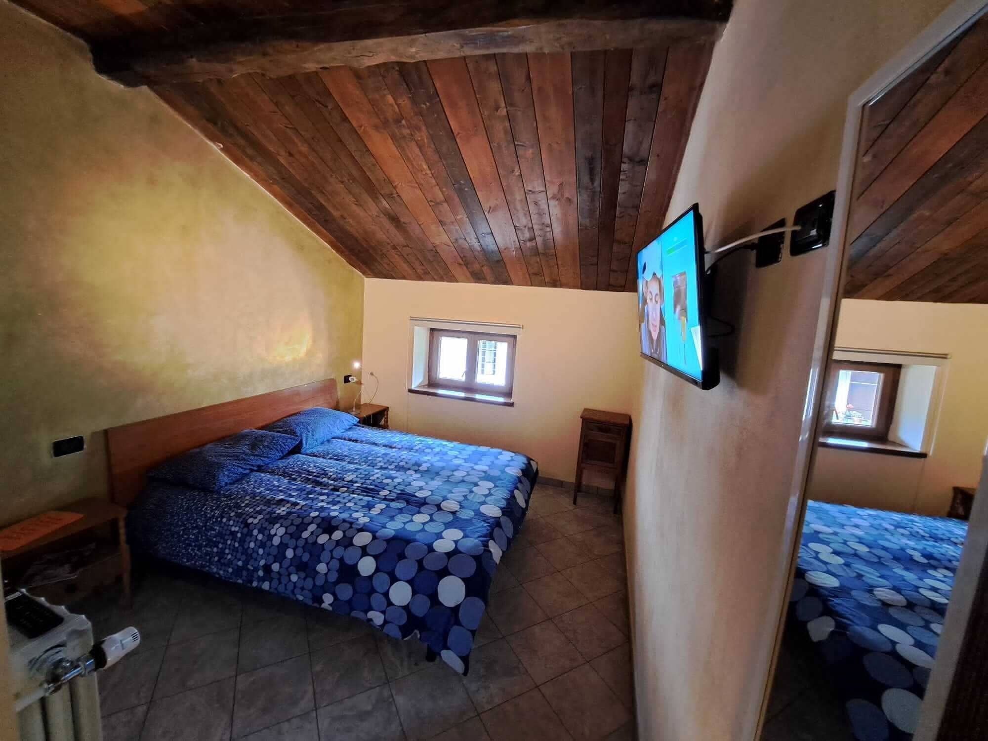 Un dormitorio con techo de madera, una cama con colcha azul de lunares, un televisor en la pared y una ventana.