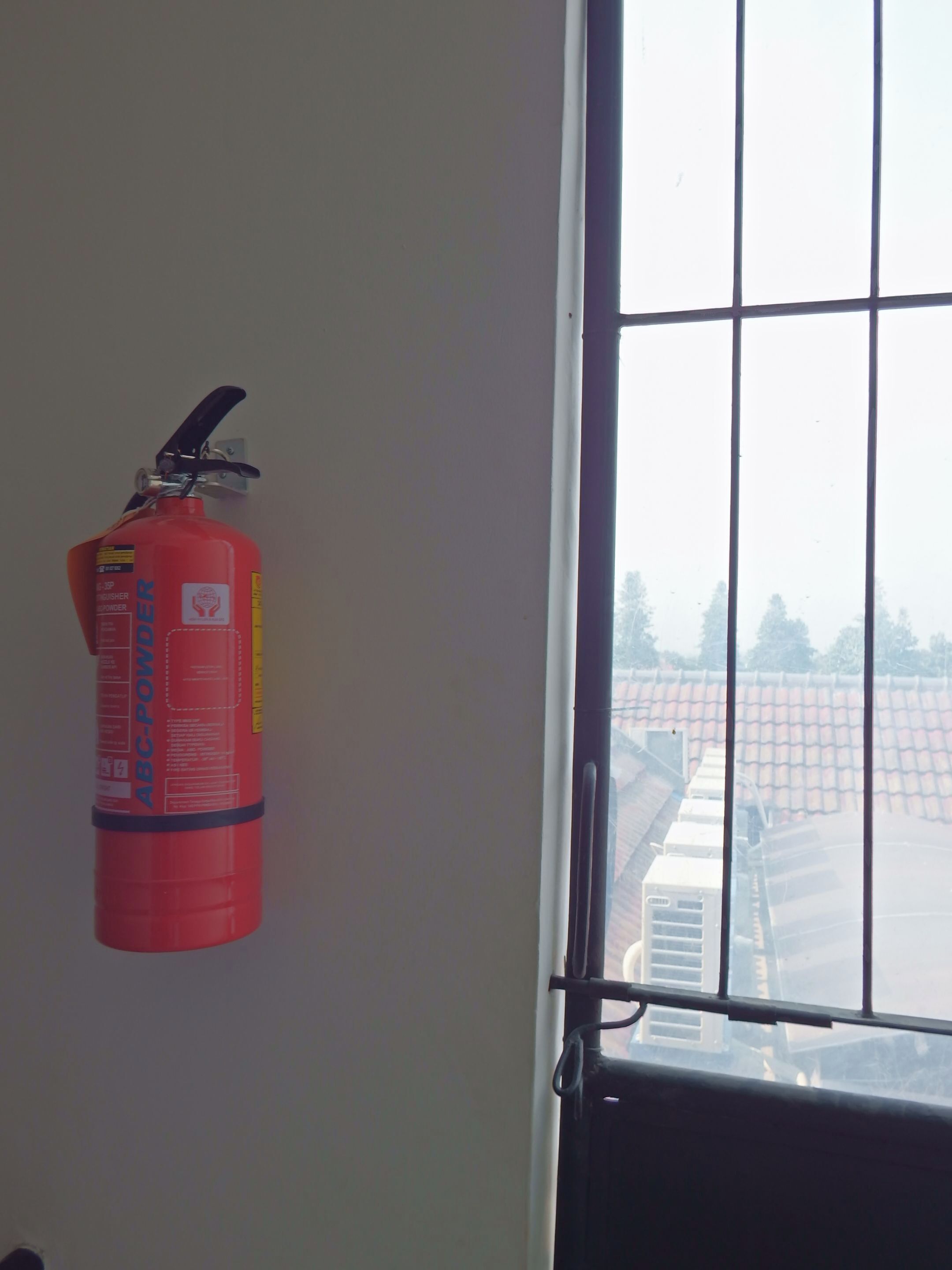 A red fire extinguisher mounted on a wall near a window with a metal grid.