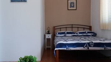A bedroom with a bed, a small bedside table, a wall-mounted air conditioner, a framed picture, and a small table with a plant.