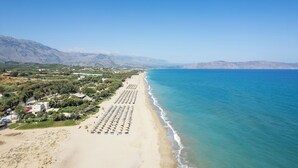 Eine Vogelperspektive zeigt einen Strand mit Reihen aus Liegestühlen und Sonnenschirmen, einem klaren, blauen Himmel und Bergen im Hintergrund.