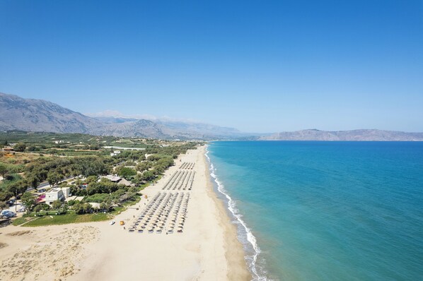 Une vue aérienne d’une plage, avec des rangées de transats et de parasols, un ciel bleu dégagé et des montagnes en arrière-plan.