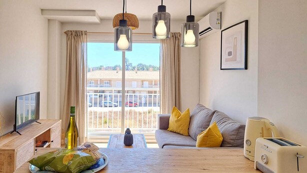 Ein modernes Wohnzimmer mit einem Holztisch, einer gelb gepolsterten Couch, einem Fernseher, einem Toaster und Blick auf eine Wohngegend durch große Fenster.